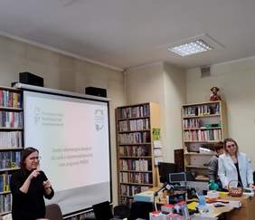 Po prawej stronie widać Panią tłumacz jezyka migowego, na środku ekran z prezentacją, a po lewej stronie Panią prelegent. W tle widoczne są regały z książkami.
