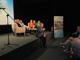 Pokaż zdjęcie: Wydarzenia na sali – osoba przemawia siedząc na scenie, obok siedzą uczestnicy spotkania, a w tle widoczny baner.