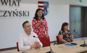 Pokaż zdjęcie: Za stołem znajdują się trzy osoby: dwie panie i pan. Kobieta w środku stoi i mówi coś do mikrofonu. Po jej bokach siedzą kobieta i mężczyzna. W tle za nimi jest napis na ścianie: Powiat Opoczyński. 