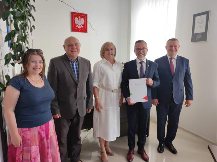Pokaż zdjęcie: Pięć osób pozujących do zdjęcia. Od lewej kobieta, mężczyzna w garniturze, za nim kobieta w długiej białej sukience i dwóch mężczyzn w garniturach. Jeden z nich prezentuje podpisaną umowę.