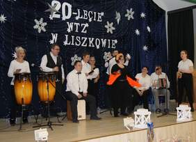 Pokaż zdjęcie: Grupa dziesięciu osób znajduje się na scenie. Większość z tych osób gra na różnych instrumentach. W tle widoczna jest granatowa kurtyna z napisem „20-lecie WTZ w Liszkowie”, która udekorowana jest gwiazdkami, śnieżynkami oraz zwisającymi lampkami.