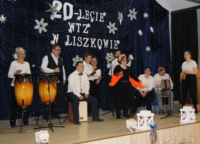 Pokaż zdjęcie: Grupa dziesięciu osób znajduje się na scenie. Większość z tych osób gra na różnych instrumentach. W tle widoczna jest granatowa kurtyna z napisem „20-lecie WTZ w Liszkowie”, która udekorowana jest gwiazdkami, śnieżynkami oraz zwisającymi lampkami.