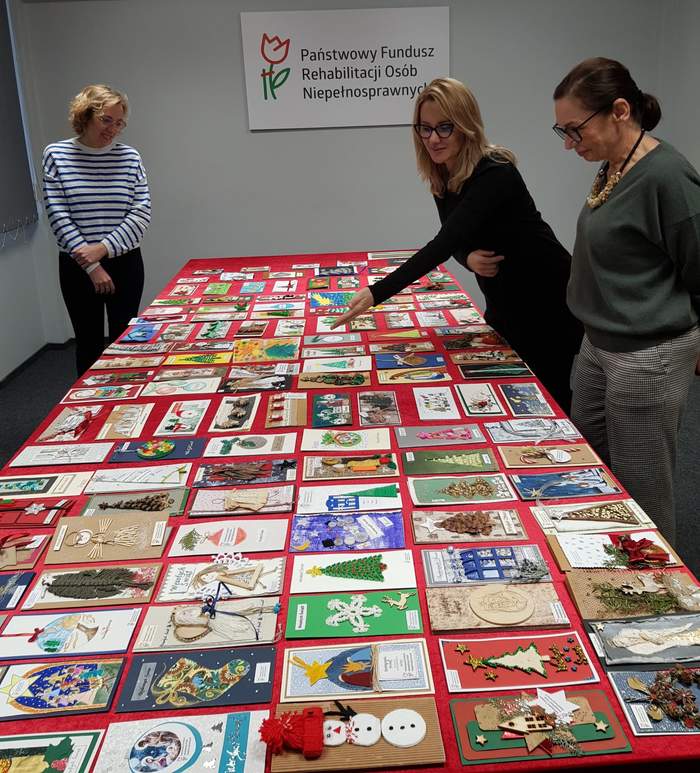 Pokaż zdjęcie: Trzy kobiety stoją wokół stołu przykrytego czerwonym płótnem i przyglądają się kartkom bożonarodzeniowym, które zostały ułożone na stole. W tle, na ścianie sali konferencyjnej widoczne jest duże logo PFRON.