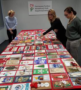 Pokaż zdjęcie: Trzy kobiety stoją wokół stołu przykrytego czerwonym płótnem i przyglądają się kartkom bożonarodzeniowym, które zostały ułożone na stole. W tle, na ścianie sali konferencyjnej widoczne jest duże logo PFRON.