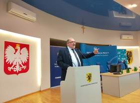 Pokaż zdjęcie: Prelegent przemawia podczas konferencji „Wizja Zero – od świadomości do działania” . Na sali widoczne są symbole województwa oraz materiały Państwowej Inspekcji Pracy.