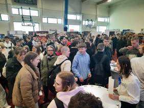Pokaż zdjęcie: W budynku szkoły na sali gimnastycznej znajduje się tłum ludzi. Są to osoby odwiedzające Targi Pracy i Edukacji. W tle widać różne stoiska wystawiennicze. 