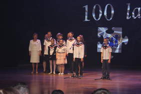 Pokaż zdjęcie: Na scenie stoją kobiety i dzieci ubrane w mundurki szkole.Wspólnie śpiewają