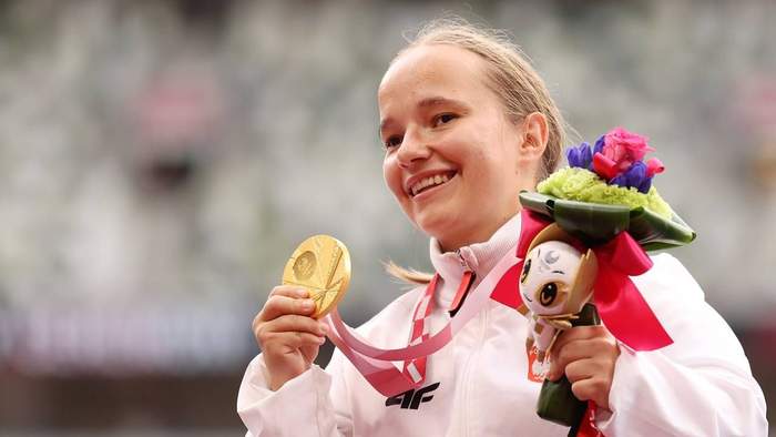 Pokaż zdjęcie: Uśmiechnięta kobieta trzyma w lewej ręce dekorację, a w prawej złotym medal Igrzysk Paraolimpisjkich