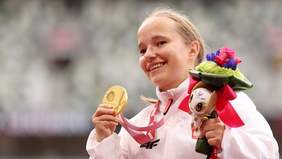 Pokaż zdjęcie: Uśmiechnięta kobieta trzyma w lewej ręce dekorację, a w prawej złotym medal Igrzysk Paraolimpisjkich