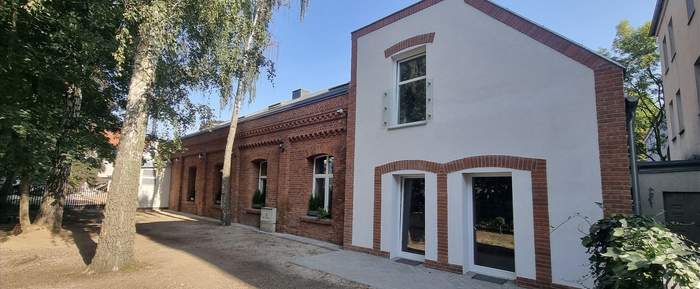 Pokaż zdjęcie: Budynek z cegły z dobudówką w białym kolorze, przed nim drzewa i trawa.
