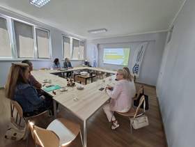 Widoczni uczestnicy spotkania siedzący przy stolikach, a w centrum wyświetlacz i prezentacja