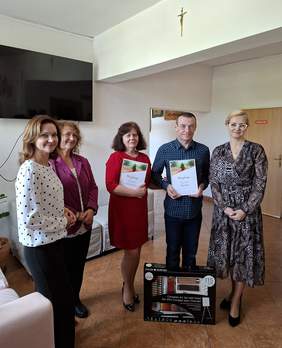 Pokaż zdjęcie: Pięć osób pozuje do wspólnego zdjęcia w pomieszczeniu WTZ. Dwie z nich trzymają dyplomy, a przed grupą stoi zestaw artystyczny jako nagroda. Na zdjęciu znajdują się przedstawiciele WTZ, TPD, PFRON oraz laureat ogólnopolskiego konkursu plastycznego.
