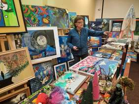 Pokaż zdjęcie: W sali znajduje sie mężczyzna. Wokół niego jest mnóstwo prac artystycznych i plastycznych. Na sztalugach wiszą obrazy. Na stole leżą rzeźby.  