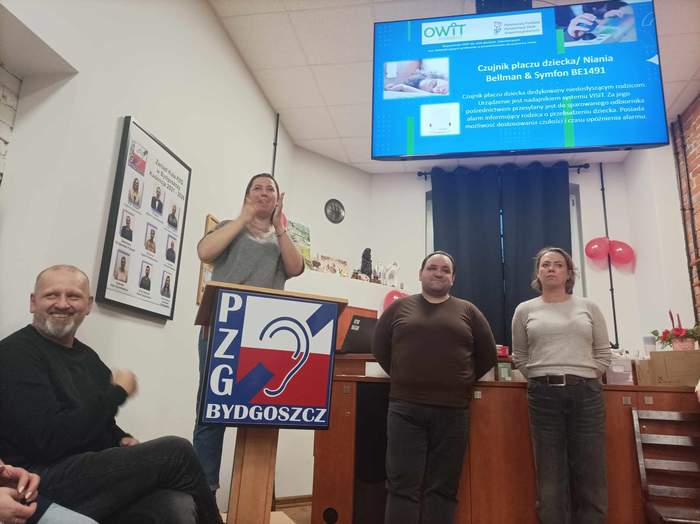 Na środku stoją trzy osoby - dwóch prelegentów i tłumaczka języka migowego, nad nimi wisi ekran z wyświetlanym slajdem