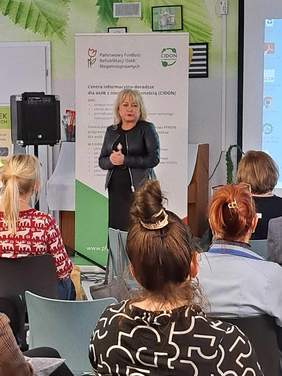 Na środku stoi Pani Róża Lewandowska - Zastępca Prezydenta Miasta Torunia przemawia, przed nią siedzą uczestnicy spotkania, a za nią stoi baner PFRON