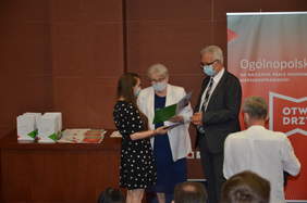 Pokaż zdjęcie: Mężczyzna i kobieta wręczają dyplom laureatce konkursu. Na ich twarzach widnieją maseczki. Z lewej strony stolik z nagrodami. Z prawej fragment banera konkursowego.