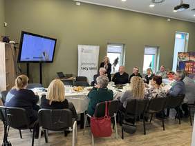 Pokaż zdjęcie: Przy stole siedzi kilkanaście osób patrzą w duży TV. Obok stoi baner z logo PFRON z napisem wszyscy jesteśmy tacy sami. Przy nim kobieta w blond włosach w czarnej marynarce.