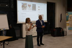 Pokaż zdjęcie: Na Sali, na szarej podłodze w duże szare marmurowe kafle stoi kobieta ubrana w zieloną suknię białą bluzkę z krótkim rękawem. Ma na sobie okulary i trzyma w ręku mikrofon. Obok stoi mężczyzna ubrany białą koszulę i granatowy garnitur. W tle