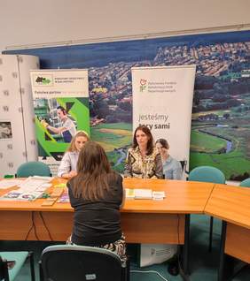 Pokaż zdjęcie: Na zdjęciu trzy kobiety siedzą przy stole, rozmawiając. W tle widoczne są banery Państwowego Urzędu Pracy w Białymstoku i Państwowego Funduszu Rehabilitacji Osób Niepełnosprawnych, oraz panorama miasta. Na stole leżą materiały informacyjne.