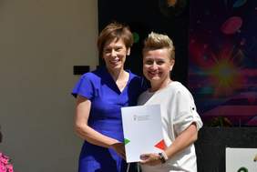 Pokaż zdjęcie: Dwie uśmiechnięte kobiety ubrane w granatową i białą sukienkę stoją obok siebie. Jedna z kobiet trzyma w dłoniach teczkę z logo PFRON.