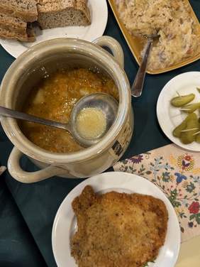 Pokaż zdjęcie: Stół z tradycyjnymi potrawami. W centralnej części znajduje się ceramiczny garnek z zupą, obok talerz z panierowanym kotletem, kromki chleba na białym talerzu, a także taca z duszoną kapustą. Na talerzyku widoczne są plasterki kiszonych ogórków.
