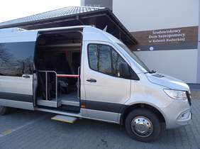 Pokaż zdjęcie: na zdjęciu  duży bus w szarym kolorze, ma otwarte drzwi gdzie można zobaczyć miejsca do siedzenia. Bus stoi na placu przed wejściem do Środowiskowego Domu Samopomocy w Kolonii Raduckiej 