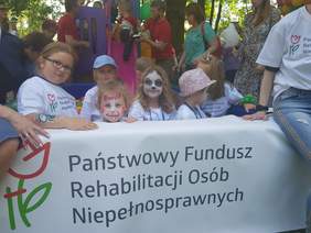 Pokaż zdjęcie: Dzieci z pomalowanymi twarzami oparte o baner z napisem Państwowy Fundusz Rehabilitacji Osób Niepełnosprawnych