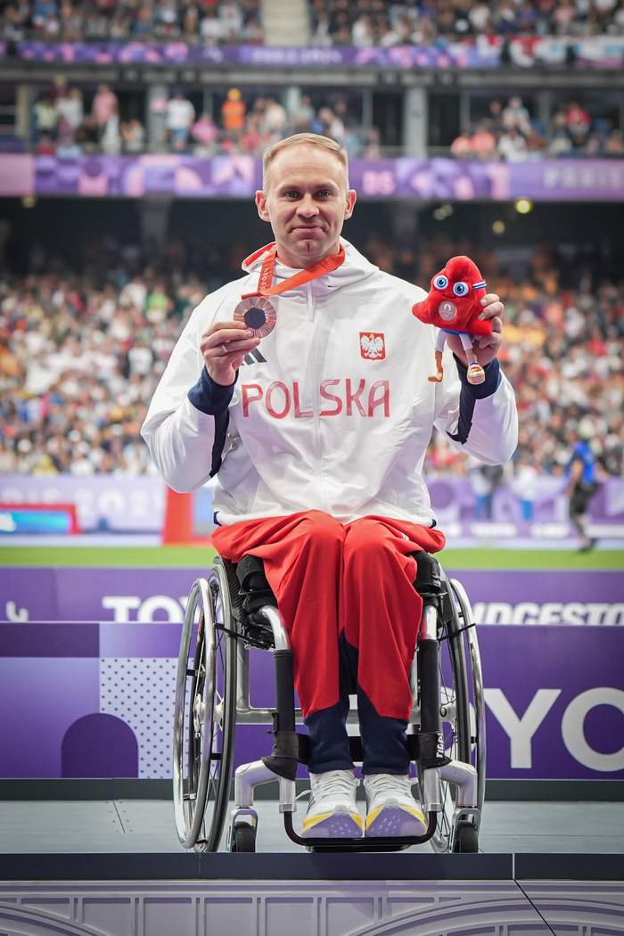 Pokaż zdjęcie: Mężczyzna na wózku prezentuje brązowym medal, jest ubrany na sportowo, w barwach reprezentacji Polski i jest uśmiechnięty