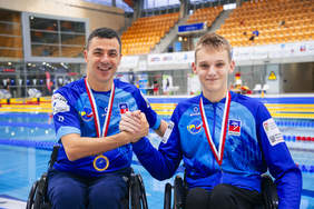 Pokaż zdjęcie: Zbliżenie dwóch uśmiechniętych mężczyzn w niebieskich kurtkach i na wózkach inwalidzkich, stojących przy basenie. Obaj mają na szyjach medale na biało-czerwonych wstążkach. Starszy mężczyzna po lewej stronie, z medalem na dodatkowej niebieskiej wstążce z logo, uśmiecha się i trzyma rękę młodszego mężczyzny po prawej, który również się uśmiecha. Na ubraniach widoczne są loga sponsorów i emblematy z napisem "Szczecin". W tle widoczne są trybuny hali basenowej.