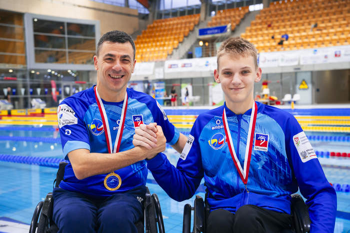 Pokaż zdjęcie: Zbliżenie dwóch uśmiechniętych mężczyzn w niebieskich kurtkach i na wózkach inwalidzkich, stojących przy basenie. Obaj mają na szyjach medale na biało-czerwonych wstążkach. Starszy mężczyzna po lewej stronie, z medalem na dodatkowej niebieskiej wstążce z logo, uśmiecha się i trzyma rękę młodszego mężczyzny po prawej, który również się uśmiecha. Na ubraniach widoczne są loga sponsorów i emblematy z napisem "Szczecin". W tle widoczne są trybuny hali basenowej.