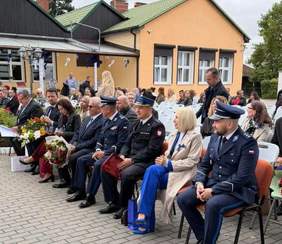 Uczestnicy uroczystości jubileuszowej 25-lecia OREW w Nowielinie siedzą przed budynkiem ośrodka. W pierwszym rzędzie widoczni są przedstawiciele służb mundurowych oraz zaproszeni goście, a wokół znajdują się pozostali uczestnicy wydarzenia.