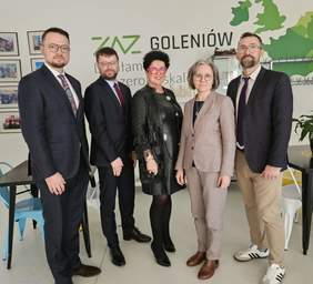 Pokaż zdjęcie: Na zdjęciu jest 5 osób, w środku dwie Panie. Pozują do zdjęcia. W tle Zielono czarny napis ZAZ Goleniów i grafika z zieloną mapą