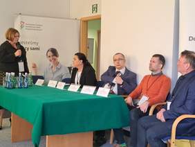 przy stole konferencyjnym siedzą prelegenci Konferencji, reprezentujący środowisko osób niepełnosprawnych oraz stoi Dyrektor Aleksandra Gierej, moderator panelu