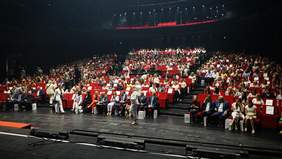 Pokaż zdjęcie: Zdjęcie sali konferencyjnej, widowni. Na widowni zasiada dużo osób, światło jest przyciemnione.