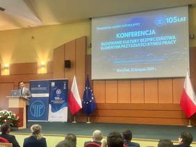 Pokaż zdjęcie: Mężczyzna w garniturze przemawia na podium podczas konferencji. W tle widoczne są slajd z tytułem wydarzenia, logo Państwowej Inspekcji Pracy, polska i unijna flaga oraz banery informacyjne. Na sali siedzą słuchacze.