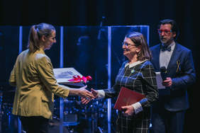 Pokaż zdjęcie: Kobieta w jasnej marynarce i spiętych ciemnych włosach, przekazuje dyplom z czerwoną kokardą kobiecie w ciemnej marynarce i okularach. Kobiety wyciągają do siebie dłonie. Za nimi stoi mężczyzna w niebieskim garniturze i w okularach.