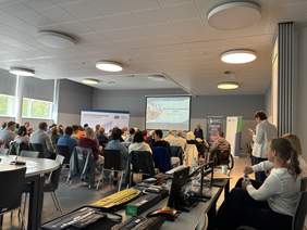 Pokaż zdjęcie: Sala konferencyjna z grupą ludzi siedzących i patrzących na wyświetlaną prezentację. Prelegent stoi z przodu przy ekranie projekcyjnym. Za prelegentem stoją pionowe roll-upy z logotypami. Na pierwszym planie widoczny jest długi stół z rozłożonym sprzętem wspomagającym i asystującym dla osób z niepełnosprawnościami.