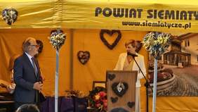 Pokaż zdjęcie: Kobieta w jasnym stroju przemawia przy mównicy pod żółtym namiotem z napisem „Powiat Siemiatycki”, obok stoi mężczyzna w garniturze. W tle dekoracje w kształcie serc i fotografia budynku DPS w Siemiatyczach.