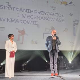 Pokaż zdjęcie: Scena w  zabytkowej Auli Akademii Sztuk Pięknych. Na scenie prowadzący wydarzenie kobieta i mężczyzna, oboje elegancko ubrani. W tle ekran, widoczny napis: Spotkanie Przyjaciół i Mecenatów ASP w Krakowie