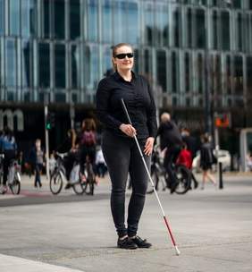 Pokaż zdjęcie: na zdjęciu osoba niewidoma poruszająca się o białej lasce, przemieszcza się wśród innych osób na ulicy