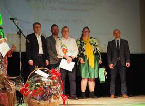 Pokaż zdjęcie: Na scenie stoją mężczyźni i kobiety - organizatorzy i laureaci konkursu. Lauretka trzyma w ręce nagrodę przekazaną przez Małopolski Oddział PFRON