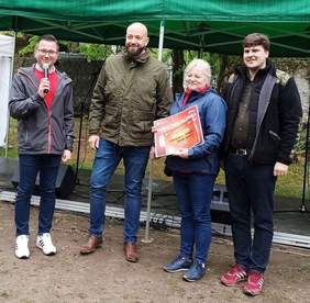 Pokaż zdjęcie: Na zdjęciu 3 mężczyzn i kobieta, która trzyma dyplom. Jeden z mężczyzn trzyma mikrofon. Są w lesie.