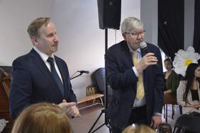 Pokaż zdjęcie: Dyrektor Łódzkiego Oddziału PFRON składa przez mikrofon świąteczne życzenia. Obok niego stoi jeszcze jeden mężczyzna. W tle widać inne osoby siedzące na krzesłach. 