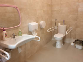 Pokaż zdjęcie: fragment łazienki wykafelkowanej na beżowo, biała umywalka i muszla klozetowa maja po bokach zamontowane uchwyty nad umywalka wisi lustro obok jest zawieszony pojemnik z papierowym ręcznikiem i elektryczna suszarka do rąk