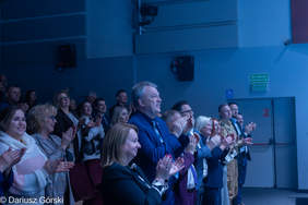Pokaż zdjęcie: Zdjęcie z widowni, przedstawiające uśmiechniętą grupę osób w eleganckich strojach, entuzjastycznie klaszczącą. W centrum kadru jest mężczyzna w niebieskim garniturze. Oświetlenie w sali jest chłodne, niebieskawe.