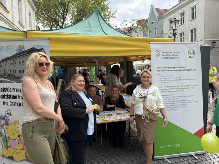 Pokaż zdjęcie: Grupa osób przy kolorowym namiocie, wśród których są osoby siedzące i stojące i widoczne są materiały promocyjne i miejsce wydarzenia