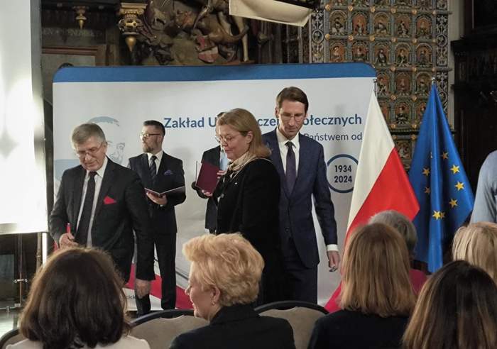 Pokaż zdjęcie: Na zdjęciu znajduje się grupa osób uczestniczących w oficjalnym wydarzeniu. W tle widoczna jest dekoracja historyczna, w tym wiszący model statku oraz zdobienia na ścianach. Na pierwszym planie widać mężczyznę w garniturze z czerwoną poszetką, obok niego stoi kobieta trzymająca jakieś dokumenty. Za nimi znajduje się baner z napisem "Zakład Ubezpieczeń Społecznych", a po prawej stronie widać flagi Polski i Unii Europejskiej. Atmosfera jest formalna, a uczestnicy są elegancko ubrani, co wskazuje na uroczysty charakter spotkania.