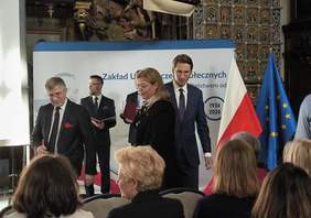 Pokaż zdjęcie: Na zdjęciu znajduje się grupa osób uczestniczących w oficjalnym wydarzeniu. W tle widoczna jest dekoracja historyczna, w tym wiszący model statku oraz zdobienia na ścianach. Na pierwszym planie widać mężczyznę w garniturze z czerwoną poszetką, obok niego stoi kobieta trzymająca jakieś dokumenty. Za nimi znajduje się baner z napisem "Zakład Ubezpieczeń Społecznych", a po prawej stronie widać flagi Polski i Unii Europejskiej. Atmosfera jest formalna, a uczestnicy są elegancko ubrani, co wskazuje na uroczysty charakter spotkania.