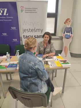 Pokaż zdjęcie: Na zdjęciu dwie kobiety siedzą przy stole, rozmawiają. W tle widoczne są banery  Państwowego Funduszu Rehabilitacji Osób Niepełnosprawnych oraz Narodowego Funduszu Zdrowia. Na stole leżą materiały informacyjne.