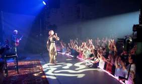 Pokaż zdjęcie: Artystka stojąca na scenie z mikrofonem w ręku przodem do stojących osób. W tle sceny widać mężczyznę z gitarą w ręku